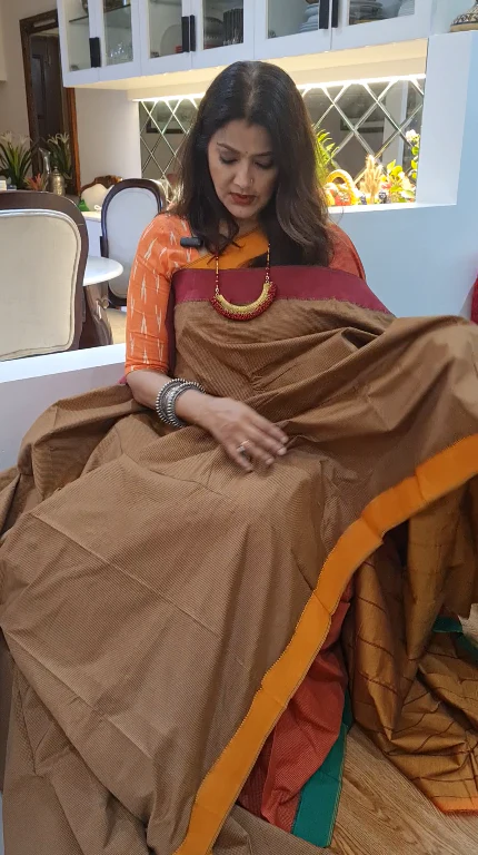 Woman in a traditional outfit with a brown saree and orange border in an indoor setting.